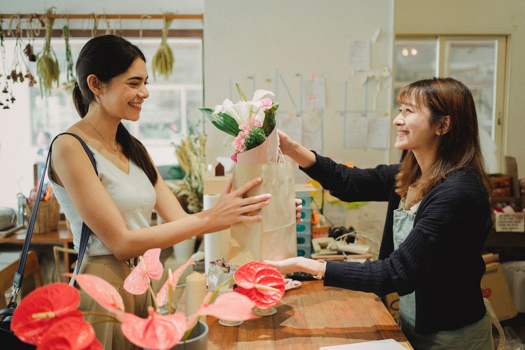 Une commerçante en Walllonie souriante fidélise une cliente avec un petit cadeau au comptoir quand elle achète un bouquet de fleurs