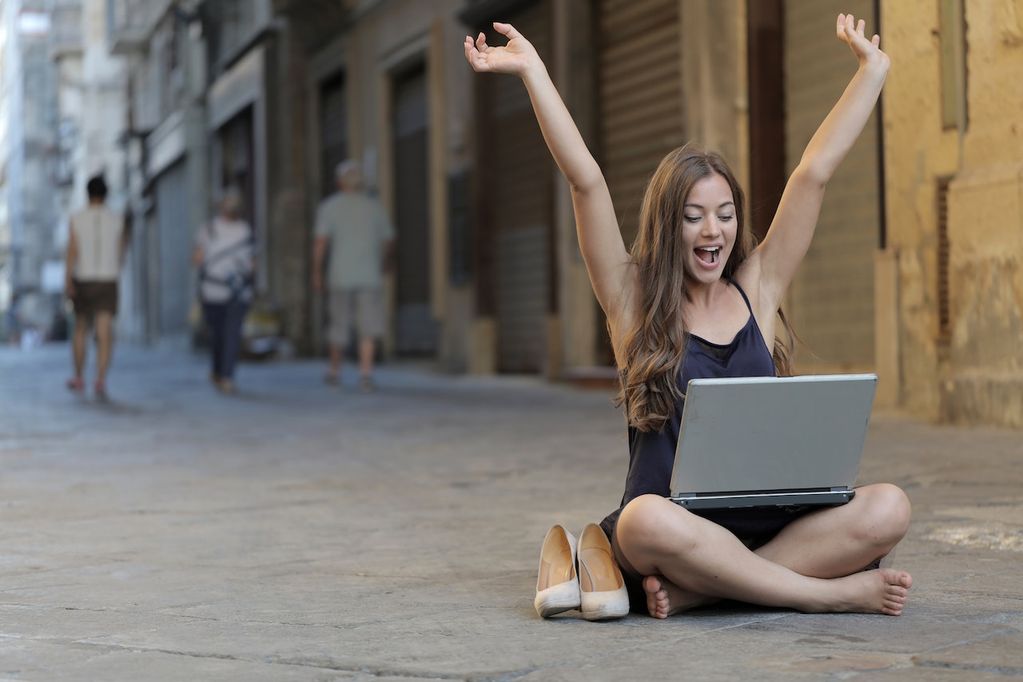 Une indépendante en Wallonie assise dans la rue lève les mains vers le ciel sur son ordinateur portable : elle a trouvé des aides et subsides pour son entreprise grâce au guide VOObusiness