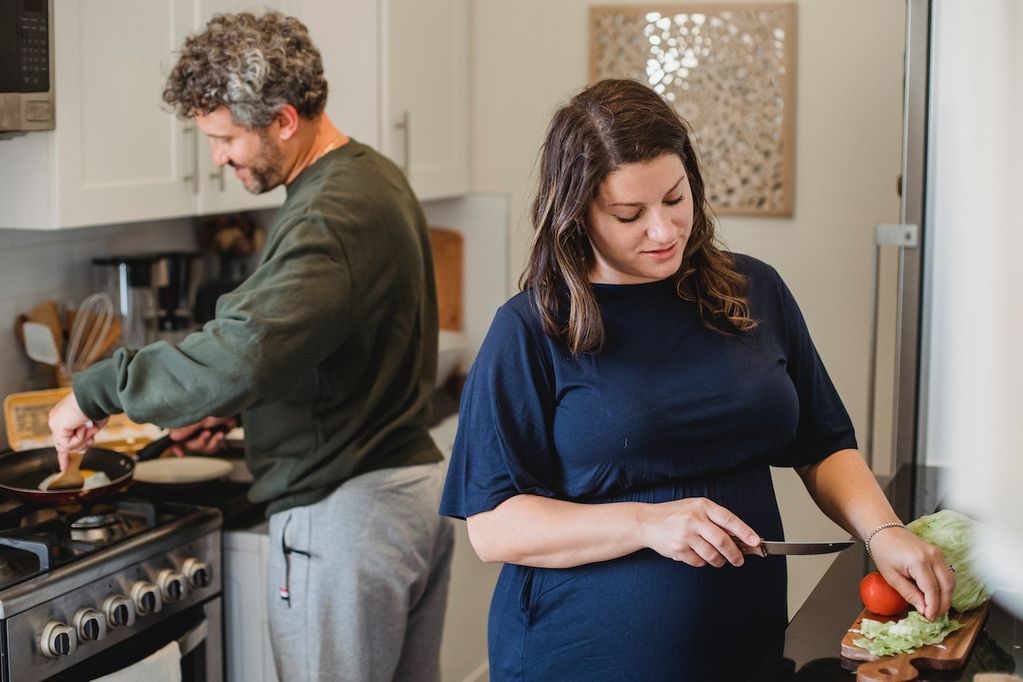 Une femme enceinte à l'avant-plan coupe des légumes dans la cuisine et son compagnon à l'arrière cuisine. C'est un couple d'indépendants en Wallonie et à Bruxelles.