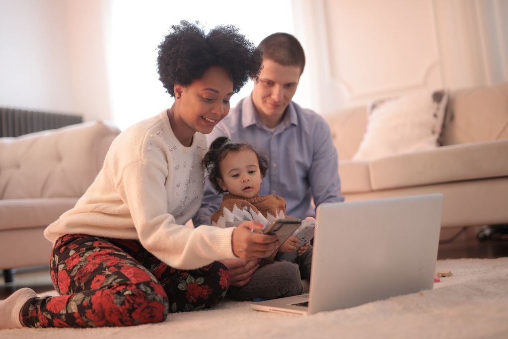 Une famille assise sur le sol une femme noire, une fille métisse et un papa caucasien en arrière plan apprennent des choses en s'amusant en famille sur le net